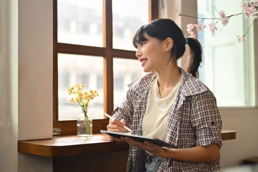 Carefree young asian woman holding digital tablet and looking away while sitting in cozy home or cafe.