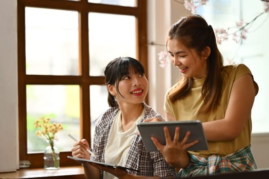 Smiling female designers holding digital tablet explaining details and sharing creative ideas to her colleague.