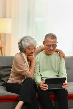 Happy senior couple dressed casually relaxing on sofa and browsing internet on digital tablet.