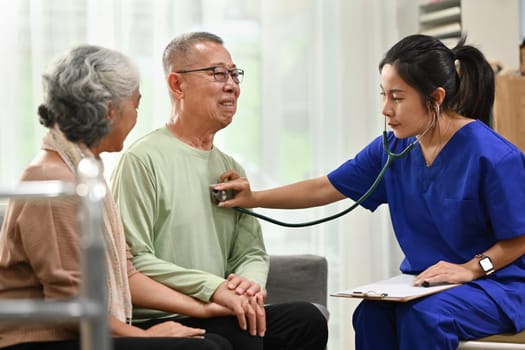 Female doctor using stethoscope examining senior male during home care visit. Elderly healthcare concept.