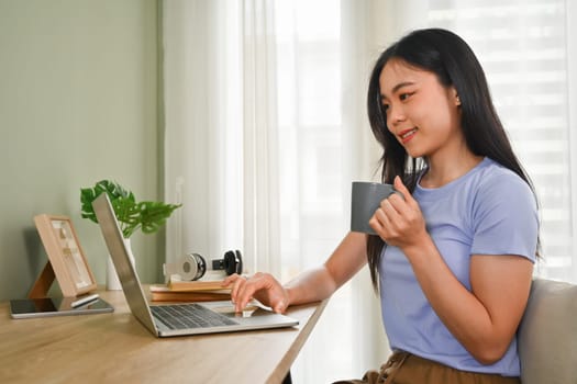 Pleased young woman in casual clothes drinking tea and reading email or news on laptop computer.