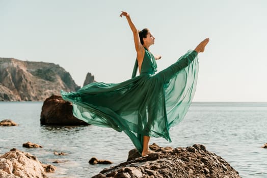 Woman green dress sea. Female dancer in a long mint dress posing on a beach with rocks on sunny day. Girl on the nature on blue sky background