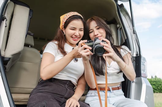 Cute Asian female friends enjoying taking photo with a camera together during a trip. and enjoy sitting and chatting in the back of the car on holiday.