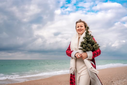 Blond woman holding Christmas tree by the sea. Christmas portrait of a happy woman walking along the beach and holding a Christmas tree in her hands. Dressed in a red coat, white dress