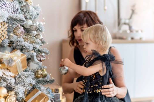 A mother with a 2 years old daughter decorates the Christmas tree. Both are dressed in black dresses, the daughter hangs a ball on the Christmas tree