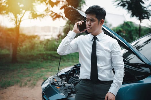 Handsome businessman in suit standing on highway, examining car engine with roadside assistance. Driver looking worried while discussing transportation problems. Serious concept