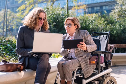 business man and woman using wheelchair working outdoors with laptop and tablet, concept of diversity and urban lifestyle