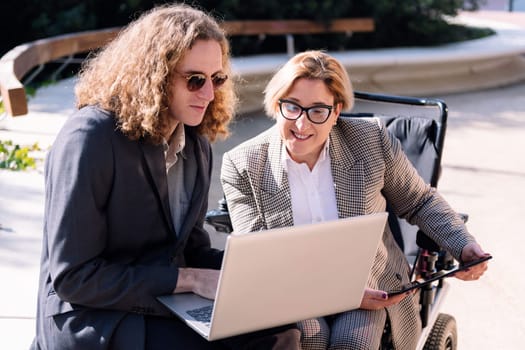 business man and smiling woman using wheelchair working outdoors with laptop and tablet, concept of diversity and urban lifestyle