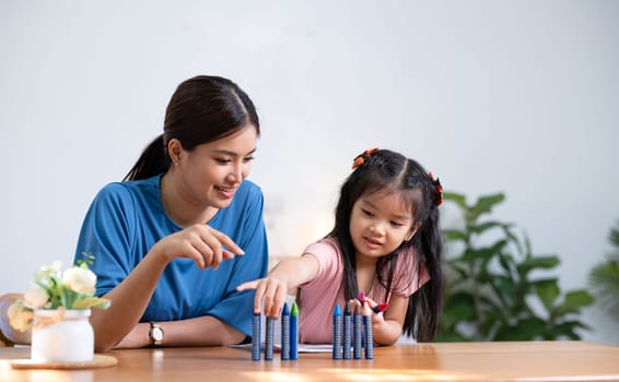 A young family spends their free time together in the living room at home. Mother and little daughter draw pictures with crayons on paper, smiling happy..