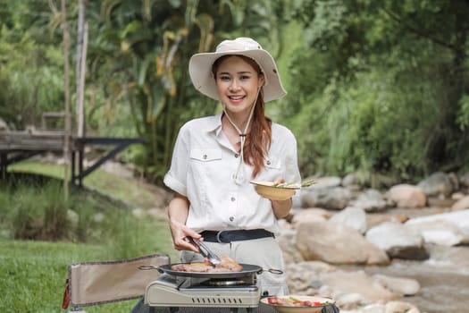 Beautiful Asian woman in her 30s preparing food Grill pork chop steak for dinner on the picnic grill. Adventure camping vacation ideas.