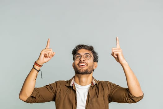 Indian bearded man showing thumbs up and pointing overhead, above head empty place, advertising area for commercial text copy space for goods promotion advertisement. Guy isolated on gray background