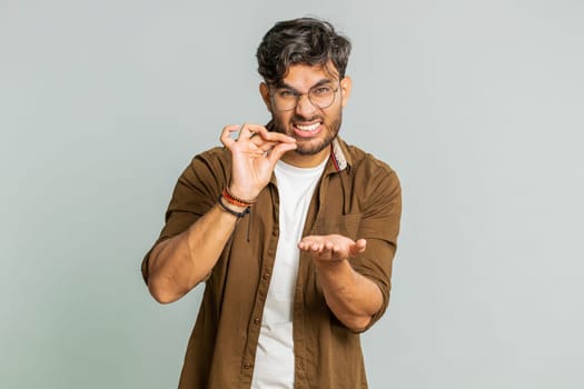 Need some more, please give me. Indian man showing a little bit gesture with sceptic smile showing minimum sign, measuring small size, begging help. Arabian guy isolated on gray studio background