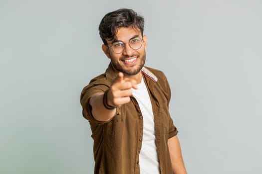 I choose you. Indian smiling man pointing finger to camera and looking with playful happy expression, making winner choice, showing direction. Arabian young guy isolated on gray studio background