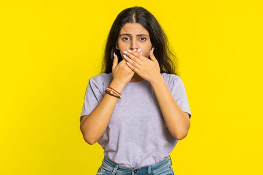 I will not say anything. Frightened Indian woman closing mouth with hands, looking intimidated scared at camera gestures no, refusing tell terrible secret unbelievable truth. Girl on yellow background