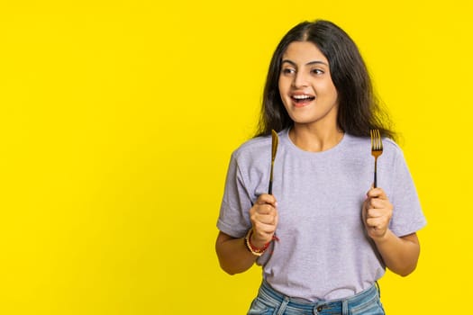 Ready to eat. Hungry Indian woman waiting for serving dinner dishes with with restlessness holding cutlery fork knife will appreciate delicious restaurant meal. Excited girl on yellow background