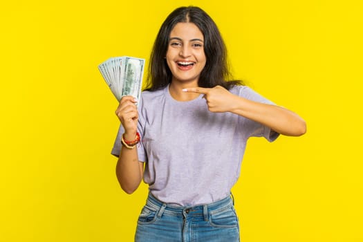 Rich pleased happy Indian young woman waving money dollar cash banknotes bills like a fan, success business career, lottery winner, big income wealth. Arabian Hindu girl isolated on yellow background