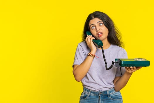 Tired bored Indian woman talking on wired vintage telephone of 80s, fooling, making silly faces, exhausted of tedious story, not interested in communication talk. Arabian girl on yellow background