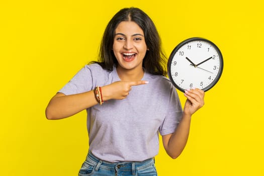 It is your time, hurry up. Indian woman showing time on wall office clock, ok, thumb up, approve, pointing finger at camera, advertisement, deadline. Arabian girl isolated on studio yellow background