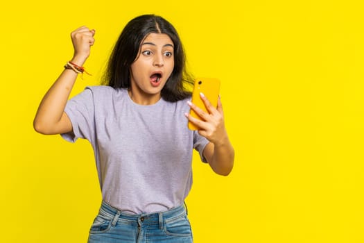 Happy excited Indian woman girl use smartphone typing browsing screaming say wow yes found out great win good news lottery goal achievement celebrating success, winning play game on yellow background