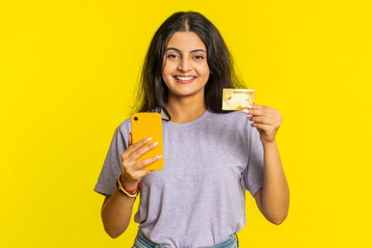 Happy Indian young woman using credit bank card and smartphone while transferring spending money, purchases online shopping, ordering food delivery. Arabian Hindu girl isolated on yellow background
