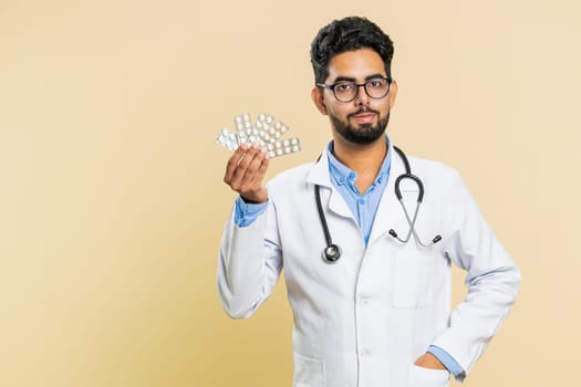 Indian doctor cardiologist man shows lot of tablet pills in blisters, recommends drugs vitamin antibiotic cure treatment. Health care. Arabian apothecary pharmacist guy isolated on beige background
