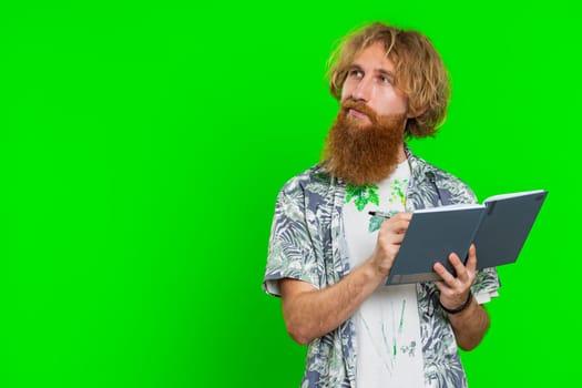 Thoughtful journalist young man making notes, writing down thoughts with pen into notepad notebook diary, to do list, good idea. Handsome redhead student guy isolated on green chroma key background