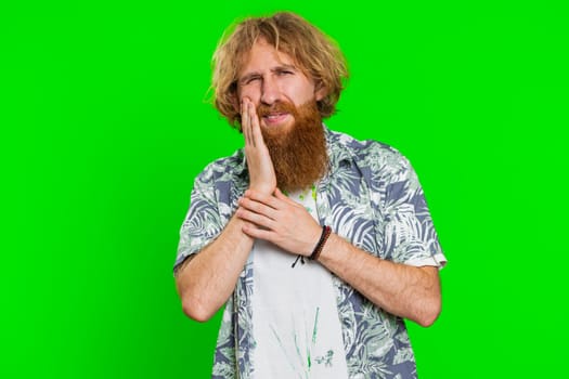 Dental problems. Young man touching cheek, closing eyes with expression of terrible suffer from painful toothache, sensitive teeth, cavities. Handsome redhead guy isolated on chroma key background