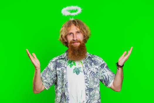 Portrait of smiling shy angelic young caucasian man with angel halo nimb over head flirting, looking at camera, positive love emotions celebrating holiday. Redhead bearded guy on chroma key background