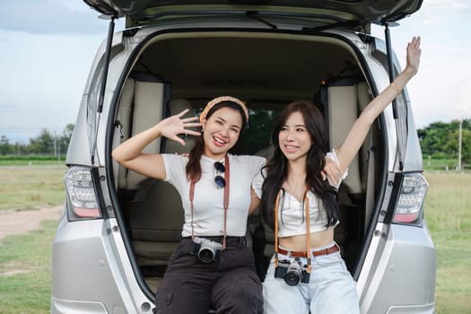 Two young Asian women in white shirts and jeans sit in the back of their new car, lounging on the field. holidays and traveling.