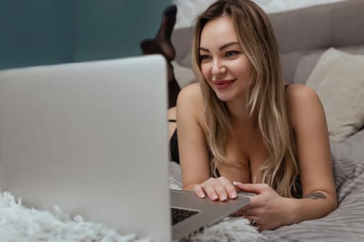 Sexy woman in black lingerie lies on the bed and uses a laptop. Sex model concept on webcam and dating site.