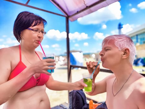 A middle-aged couple shares a joyful moment on the beach, sipping colorful drinks under a bright sun. Their playful connection highlights their friendship and love during vacation.