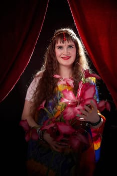 A mature woman poses gracefully in a colorful sari, holding blooming flowers, while celebrating Indian traditions in an elegant studio setting.