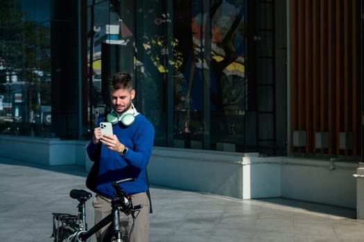 Young caucasian man using smartphone while standing next to his bicycle in the city. Sustainable commuting concept.