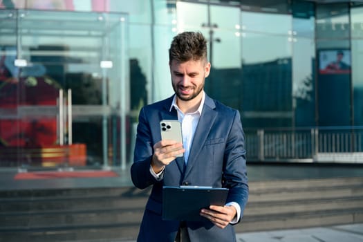 Confident businessman checking message on smartphone, standing outside an office building.