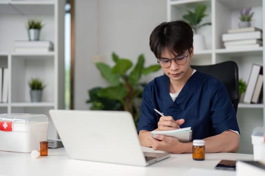 A focused medical student in scrubs takes notes while studying at a desk, surrounded by a laptop and medication bottles, showcasing dedication to learning.