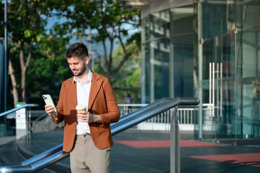 Caucasian businessman holding smartphone and a takeaway coffee cup standing outside office building.