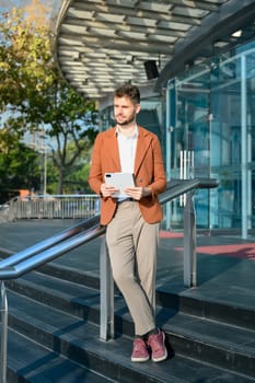 Portrait of successful businessman holding digital tablet leaning on a railing outside a glass office building.
