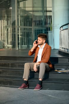 Caucasian male entrepreneur talking on phone sitting on the steps outside an office building.