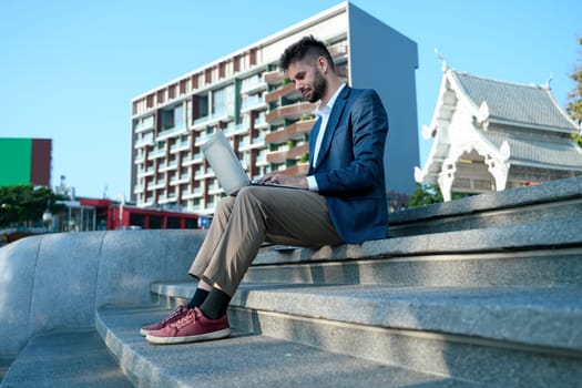 Handsome caucasian businessman sitting on urban steps working on his laptop.