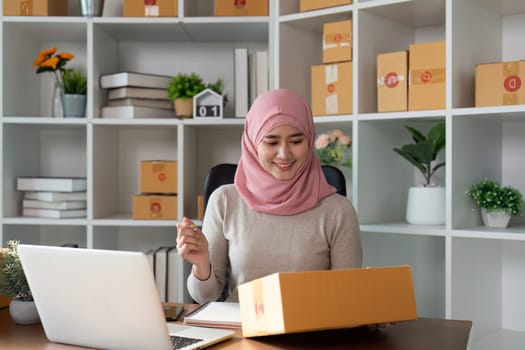 A Muslim woman in a hijab conducts online sales and manages her business efficiently from a contemporary workspace with packages around her.