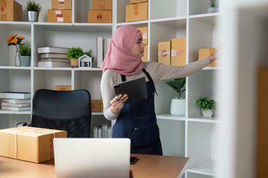 A Muslim entrepreneur in a hijab organizes online orders and manages her business using a tablet in a stylish office setting.