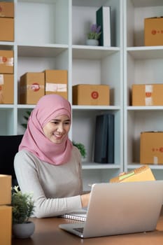 A cheerful Muslim woman in a hijab manages her online business at a computer, showcasing her entrepreneurial spirit in a contemporary workspace.