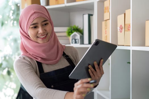 A Muslim woman in a hijab uses a tablet to manage her online sales and inventory, showcasing entrepreneurship and digital commerce.