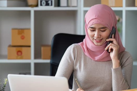 A Muslim woman in a pink hijab engages in online sales, managing orders and customer communication in her office.