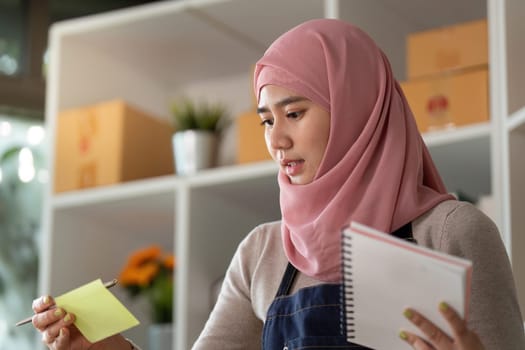 A focused Muslim woman in a hijab reviews notes while managing her online sales, showcasing entrepreneurship and dedication.