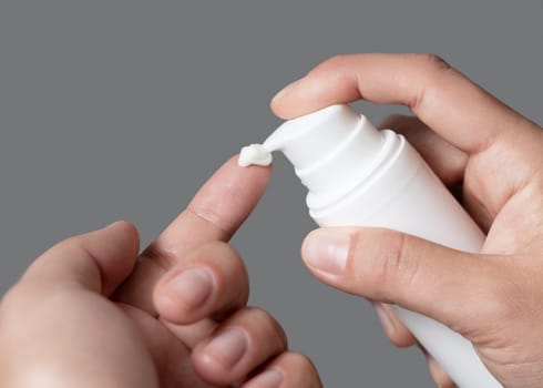 Close up of woman squeezing hand cream on her finger and holding a cosmetic bottle in other hand against grey. Beauty treatment or handcare procedure in a spa salon