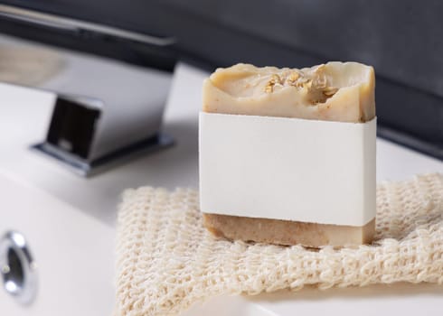 Beige handmade soap bar with blank label on sisal soap saver bag near modern faucet on basin in dark grey masculine bathroom close up, mockup. Natural herbal product for daily hygiene and bodycare