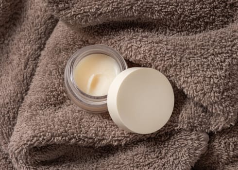 Opened lip balm jar with blank lid on light brown bath towel top view, cosmetic packaging mockup, copy space. Lipcare beauty product for mens and woman daily lipcare and personal care