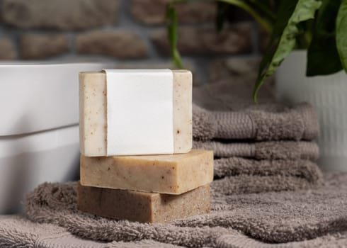 Soap bar with blank label on light brown folded towels, near green plant and vessel basin in bathroom, close up, brand packaging mockup. Men hygiene product