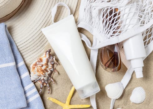 White blank cream tube, bag, towel, starfishes and seashells on a sandy beach, top view. Sunscreen cosmetics mockup. Summer vacation with spf uv-protected sunbathing